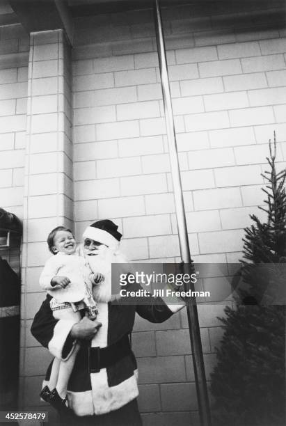 Man dressed as Santa Claus carries a young girl to the fireman's pole in a firehouse, New York City, circa 1973.
