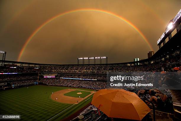 Fans sit under the cover of an umbrella as a rainbow illuminates the field while starting pitcher Brett Anderson of the Colorado Rockies delivers to...