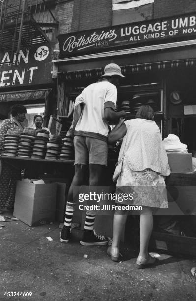 Woman holds the hand of a tall man as he browses the hats on sale at a market stall, Lower East Side, New York City, 1975.