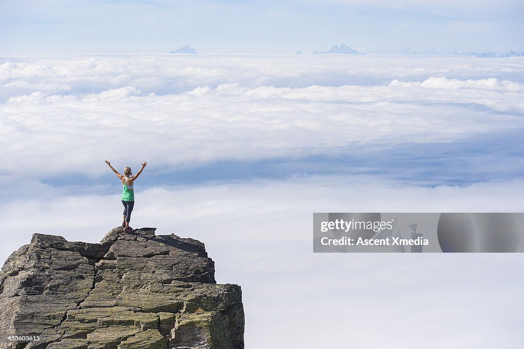 Woman stands on mt summit above clouds, arms out