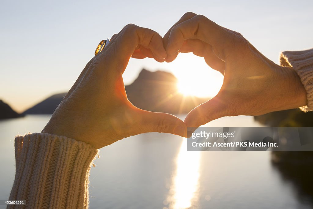 Woman's hands create heart shape at lake, sunrise