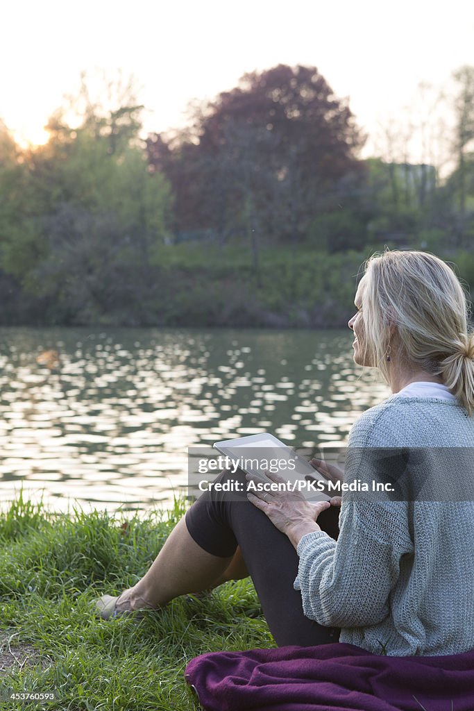 Woman relaxing by river's edge with digital tablet
