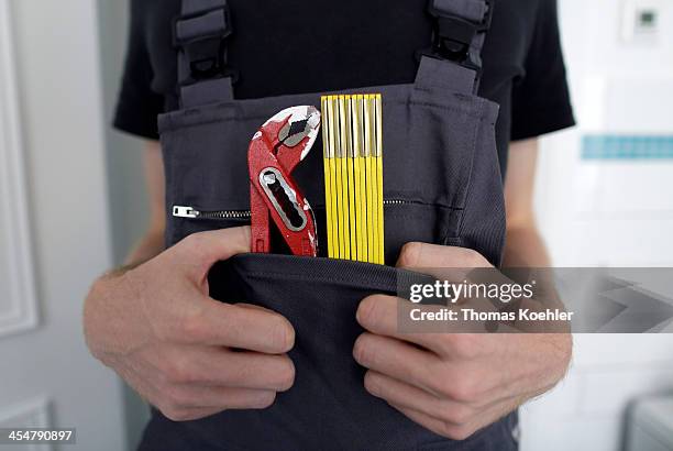 Staged picture of a pipe wrench, a folding rule and the workman's thumbs tucked into the bib pocket of a pair of dungarees he is wearing, pictured on...