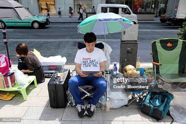 Takuya Kajigaya uses an iPad while sitting next to a mask resembling the late Apple Inc. Co-founder Steve Jobs as he waits in line outside the Apple...