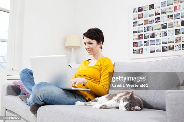 germany, bavaria, munich, mid adult woman using laptop besides cat on couch, smiling - cat laptop stockfoto's en -beelden