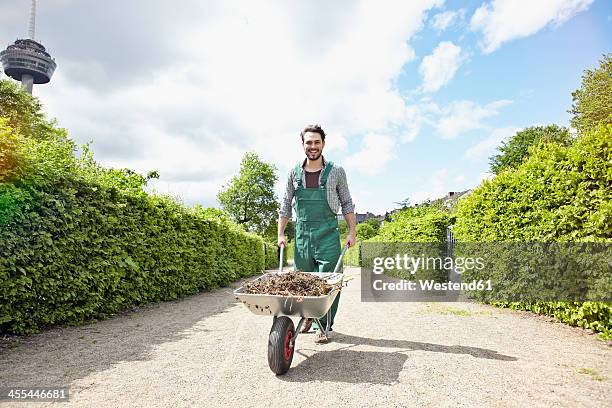 germany, cologne, portrait of young man holding wheelbarrow, smiling - tuinbroek stockfoto's en -beelden