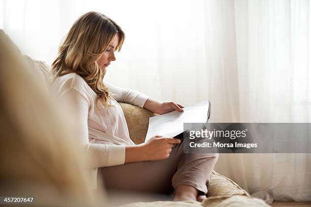 woman sitting on sofa reading magazine - reading stockfoto's en -beelden