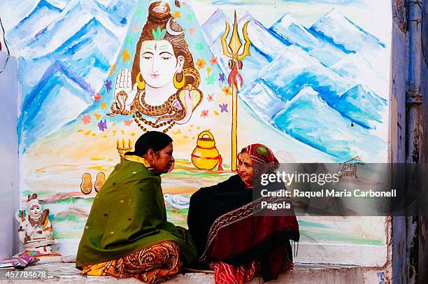 Two women sitting and talking on the street in front of a wall painted with a Shiva god image.