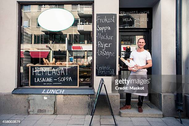 portrait of young male owner holding bread loafs while standing outside bakery - bakery shop stock pictures, royalty-free photos & images