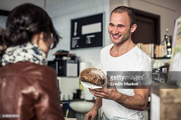 happy young male owner giving bread loaf to customer in bakery - bakery shop stock-fotos und bilder