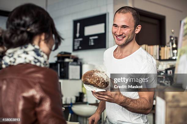 happy young male owner giving bread loaf to customer in bakery - baker stock pictures, royalty-free photos & images