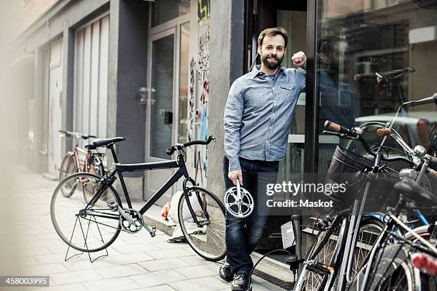 portrait of confident mid adult male owner with pedal standing at the entrance of bicycle repair shop - bike shop stock pictures, royalty-free photos & images