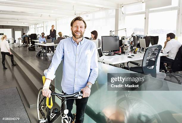 portrait of mid adult businessman holding bicycle with colleagues working in background at office - indian corporate women background stockfoto's en -beelden