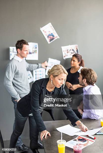 mid adult businesswoman looking at paper at desk with colleagues discussing in background - indian corporate women background stockfoto's en -beelden