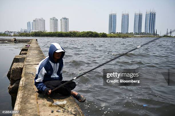 Man sits on a low sea wall while fishing as the Pantai Mutiara, rear left, and the Regatta residential projects, both developed by PT Intiland...