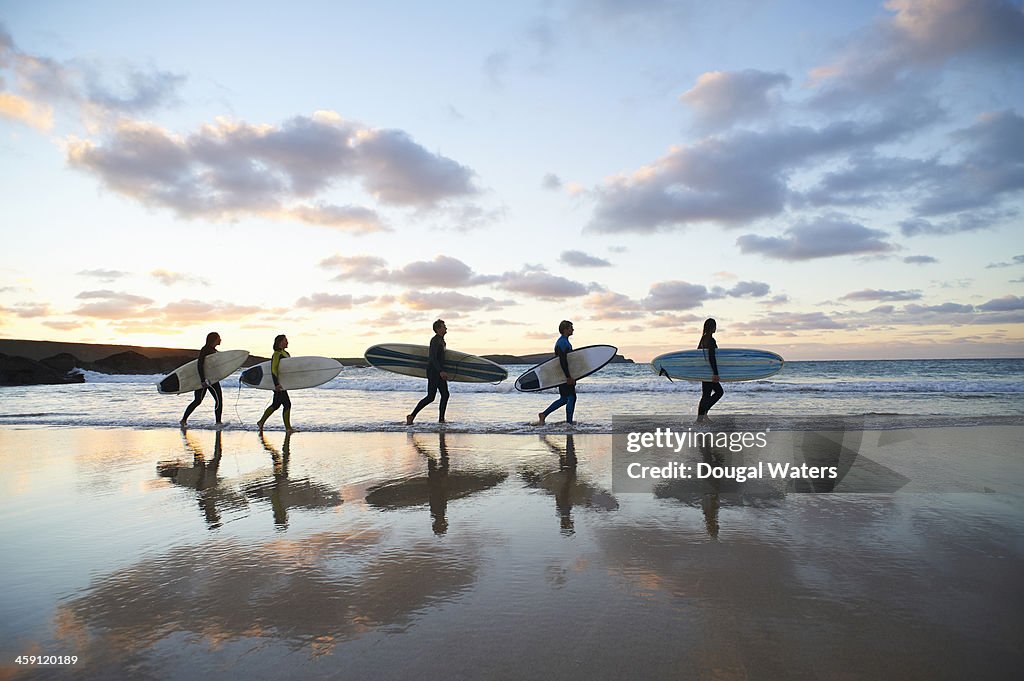 Five surfers walk along beach with surf boards.