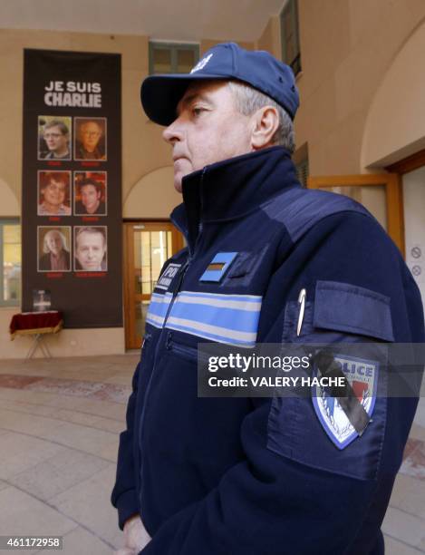 French municipal police officer with a black ribbon covering his badge stands next to a banner reading "Je suis Charlie" featuring the portraits of...