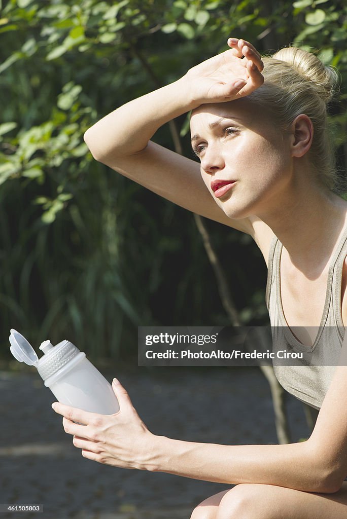 Young woman sitting outdoors with water bottle, wiping forehead with back of hand