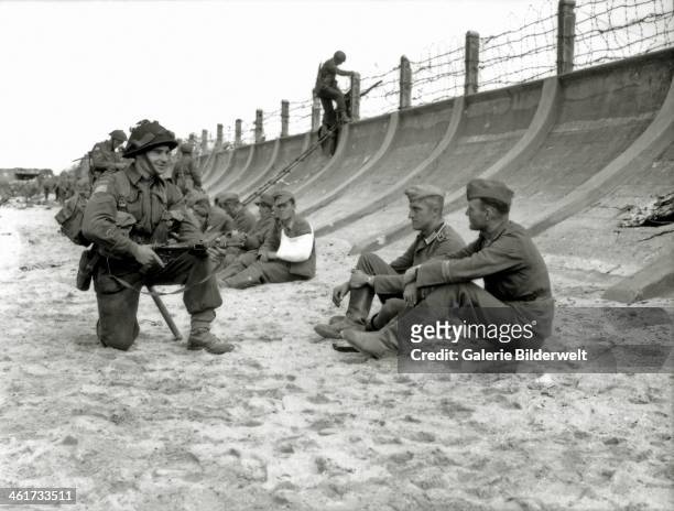 Corporal Victor Deblois from the 3rd Canadian Infantry Division crouching weapon in hand before two German prisoners sitting at the foot of the...