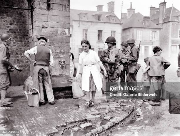 Group of American soldiers is standing at the village fountain. 12th June 1944. A woman is walking away with two pitchers while three children are...