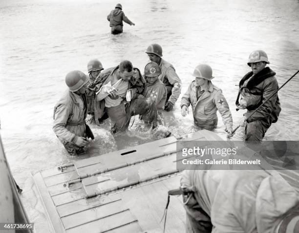 Medics of the 2nd Naval Beach Battalion help an injured paratrooper to board a Landing Craft, Vehicle, Personnel . Some medics wear a red cross on a...