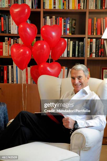 The Italian journalist Carlo Rossella poses smiling in his home with a book and some heart-shaped baloons . Pavia, 9th March 2012