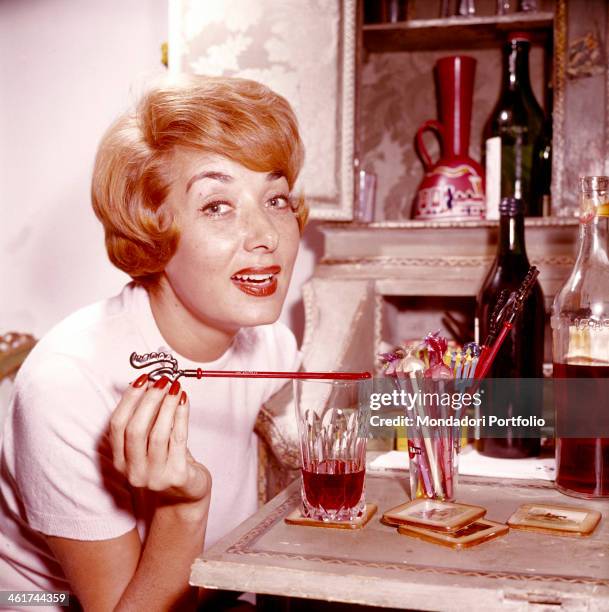 Smart Italian actress Lauretta Masiero glances seductive at the camera, with a cocktail decorations in her fingers; on the table in front of her,...