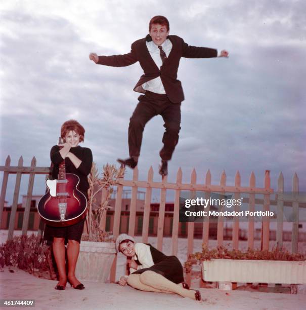 The Italian singer Tony Renis, born Elio Cesari, is jumping on a wooden fence on a beach in Sanremo; the Italian actress Laura Efrikian is curling up...