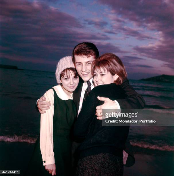 The Italian singer Tony Renis, born Elio Cesari, is smiling while embracing the Italian actresses Vicky Ludovisi and Laura Efrikian by the sea; in...