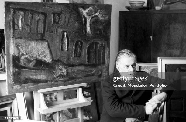 Italian painter Mario Sironi looking far seated in his atelier. Behind him, one of his Composizione di medie dimensioni standing on an easel. Italy,...