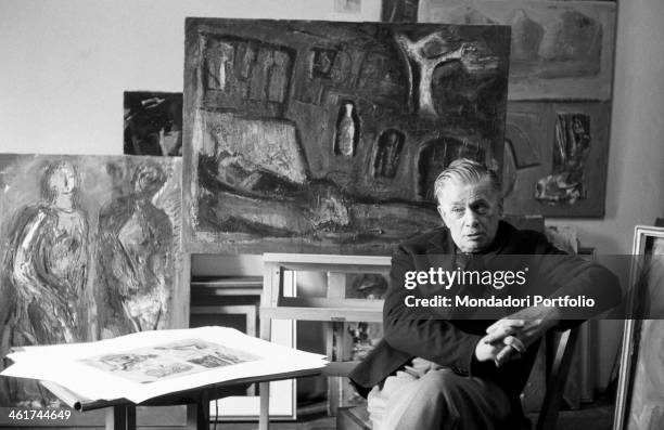 Italian painter Mario Sironi sitting in his atelier. Behind him, some of his paintings and among them one of his Composizione and a painting...