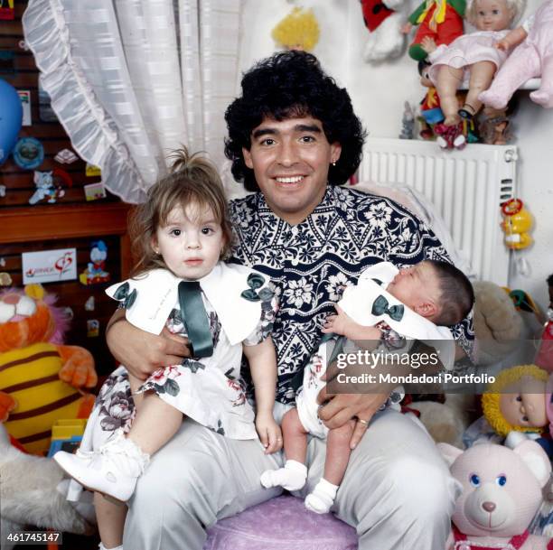 Argentinian football player Diego Armando Maradona sitting in a small bedroom with his daughters Dalma e Giannina in his arms. Italy, 1989