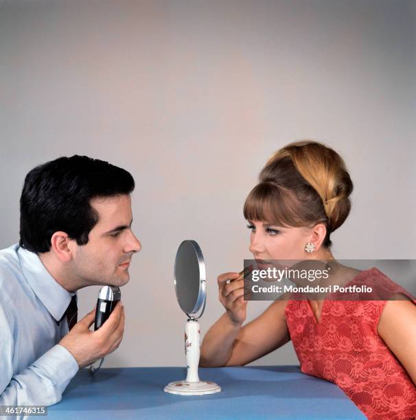 Man shaving with an electric razor in front of a double-faced mirror while a woman on the other side putting lipstick on. Italy, 1966