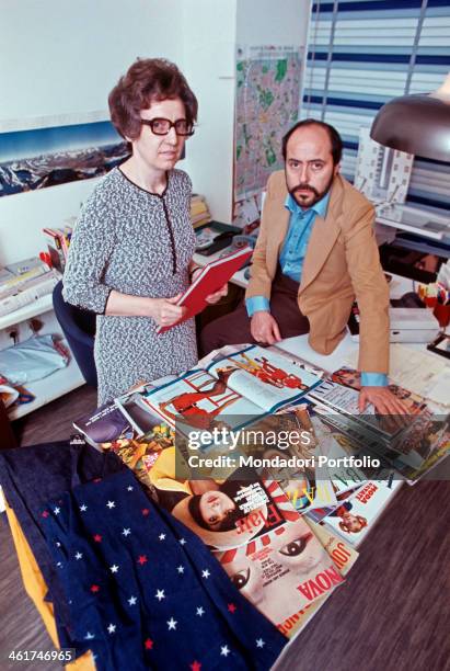 Italian fashion designer and entrepreneur Elio Fiorucci posing in his office with his secretary. On the desk, fashion magazines and fabrics. Milan,...