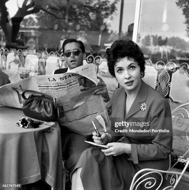 Italian actress Gina Lollobrigida sitting at the table of an outdoor bar with her husband, Slovenian doctor Milko Skofic, reading the newspaper....