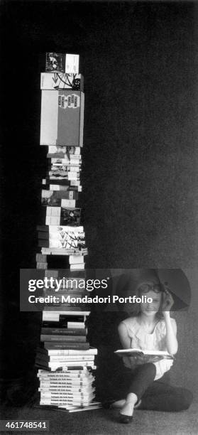 Young woman is seated on the ground beside a tall pile of volumes, with an open book on her knee, and smiles at the camera with a knowing expression,...