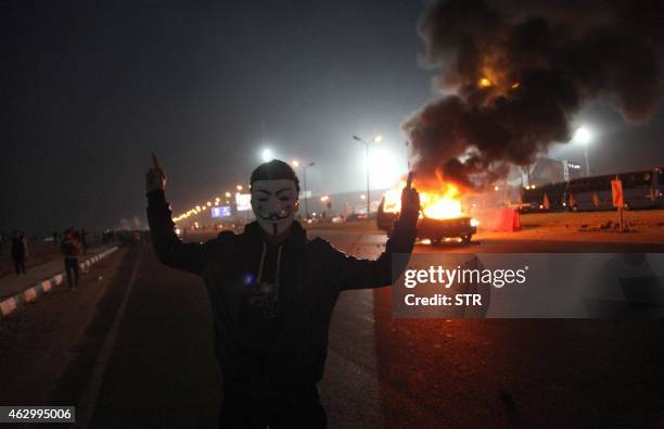 An Egyptian man wearing a mask of the anonymous movement gestures near a burning car outside a sports stadium in a Cairo's northeast district, on...