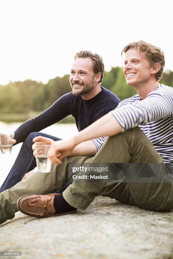 Happy male friends holding glasses of water while sitting on rock