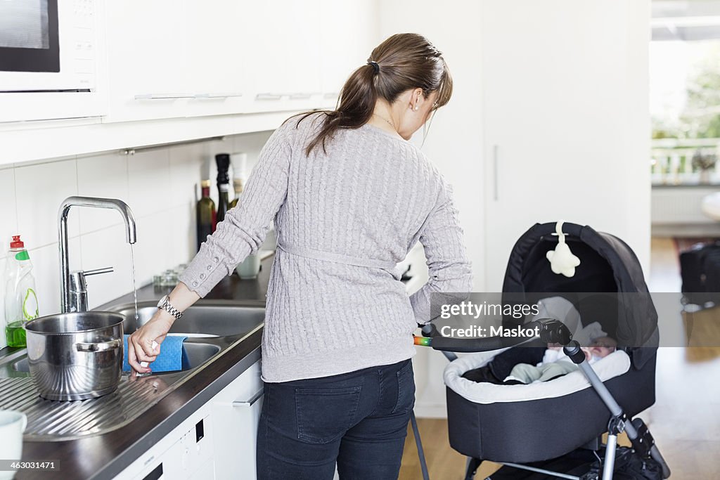 Rear view of woman looking at baby girl in carriage while washing utensils in kitchen