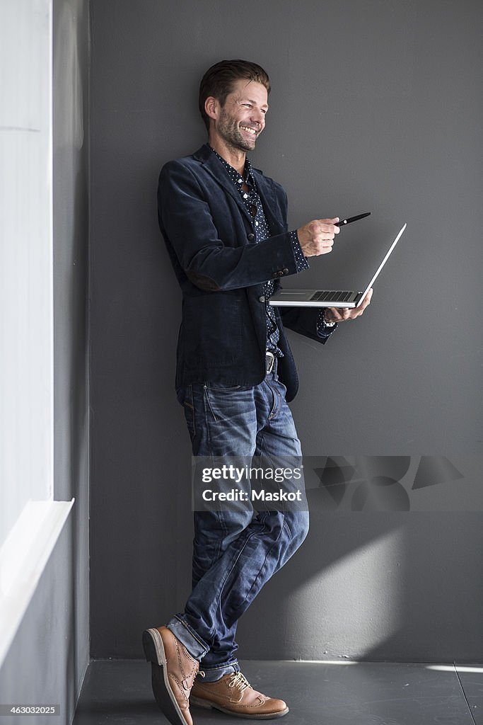 Happy businessman gesturing with pen while holding laptop against wall