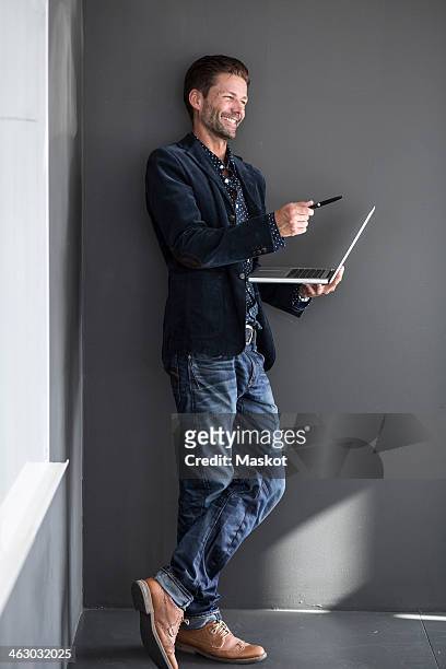 happy businessman gesturing with pen while holding laptop against wall - leaning 個照片及圖片檔