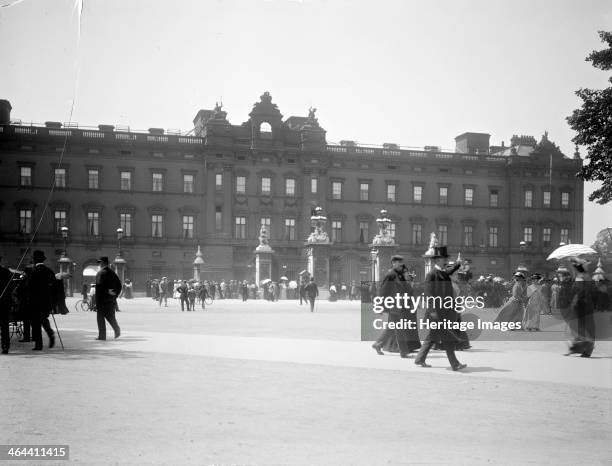 Sightseers crowd around the gates of Buckingham Palace, London. This view shows the public east facade as it looked after Edward Blore closed off the...