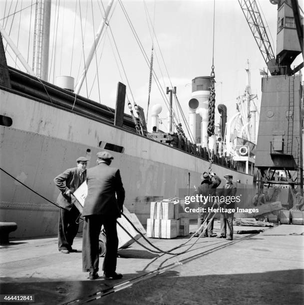 Loading a ship at the North Quay, West India Docks, London, c1945-c1965. Dock workers handle crates on the quayside for loading onto a Hellenic Lines...