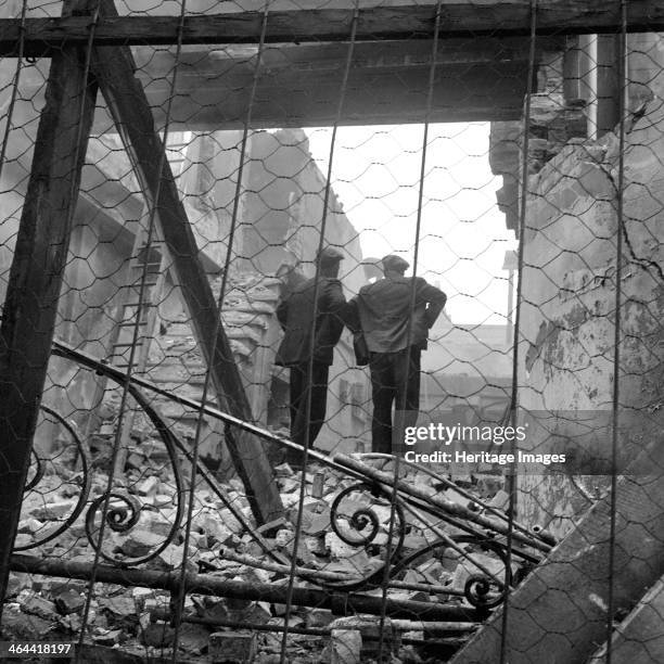 Demolition site, London, 1960-1965. Looking through chicken wire and past an ornate piece of wrought ironwork to two workmen standing on the rubble...