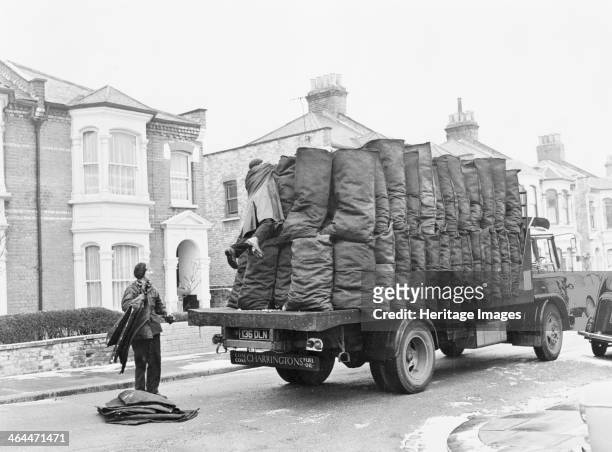 Charringtons delivering sacks of coal down a residential street. A coalman watches his colleague reaching over the sacks of coal on the back of the...