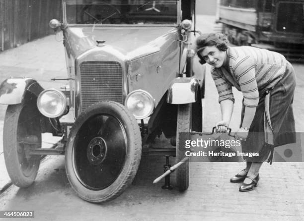Ivy Cummings changing a tyre on a 1925 Singer 10/26, London, c1925. The car is jacked up; Ms Cummings uses a spanner to tighten the bolts on the new...