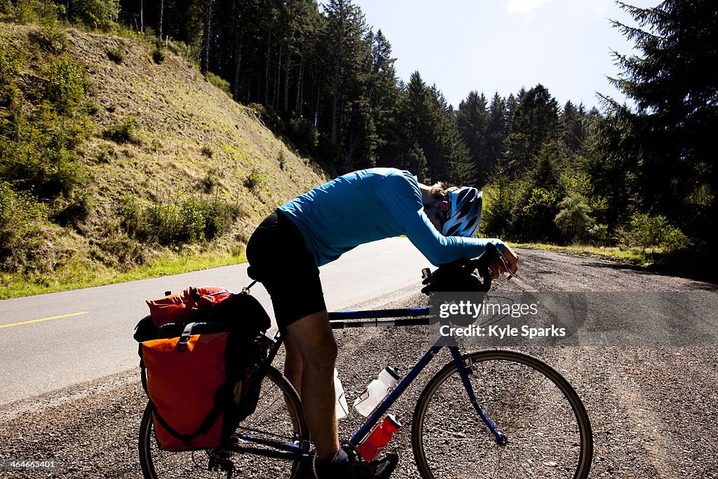 An exhausted male cyclist leans over his touring bike while climbing Mattole Road near Ferndale, California.