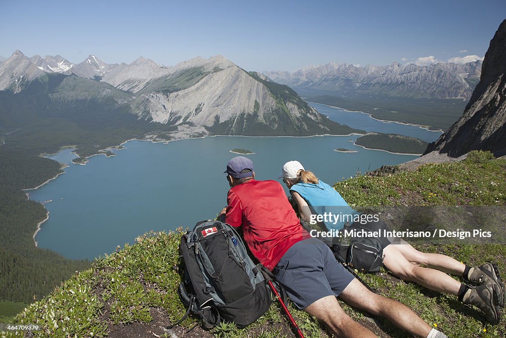 Male And Female Hiker Laying Down On A Mountain Ridge Overlooking An Emerald Lake And Mountains Below With Blue Sky In Kananaskis Provincial Park