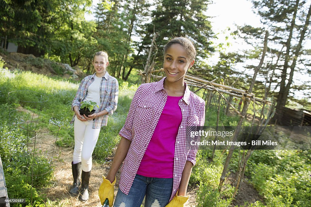 Two girls in a garden, walking up the path, one carrying a plant in a pot.