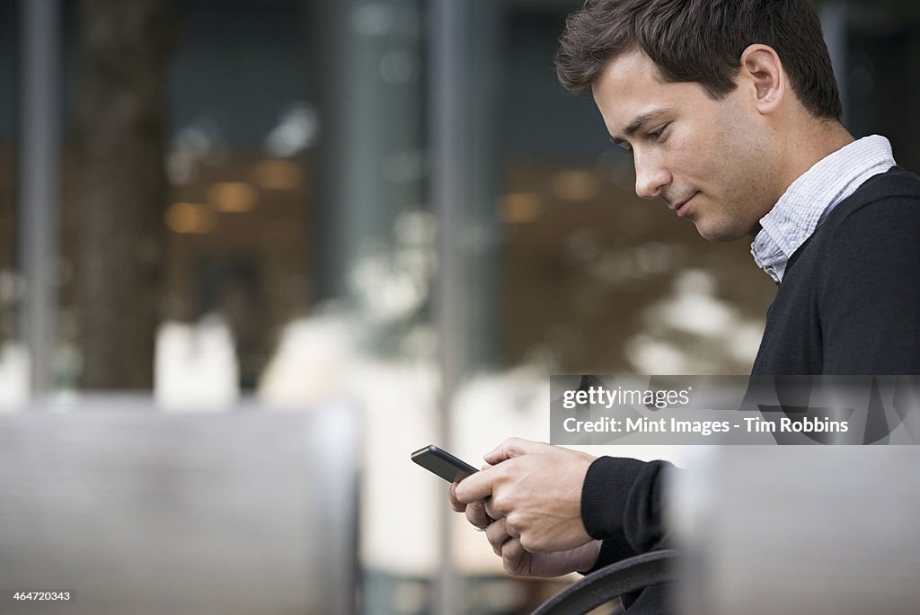 Summer in the city. People outdoors, keeping in touch while on the move. A man sitting on a bench using a smart phone.